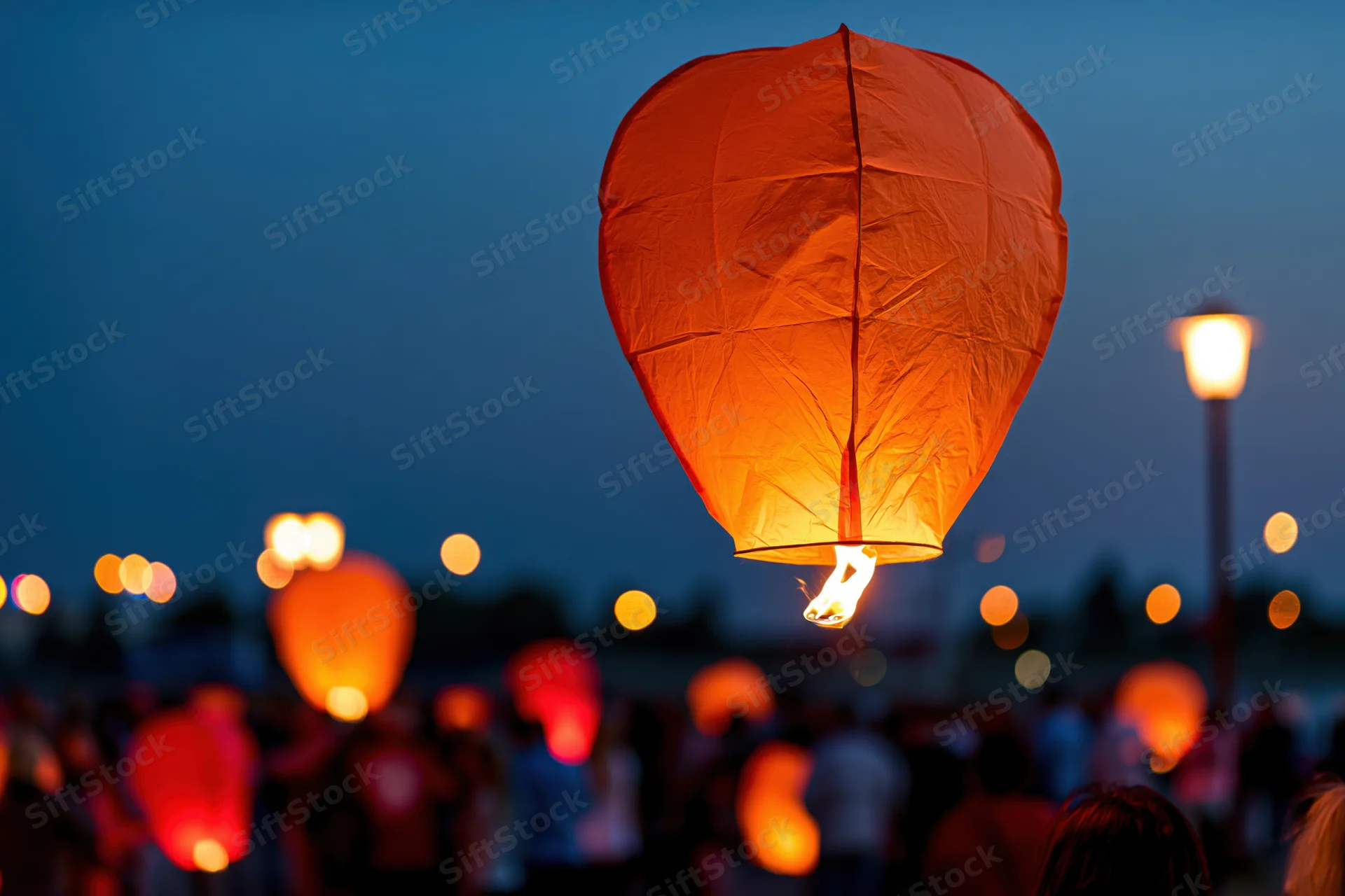 Glowing orange paper sky lantern floating upward at dusk with crowd below