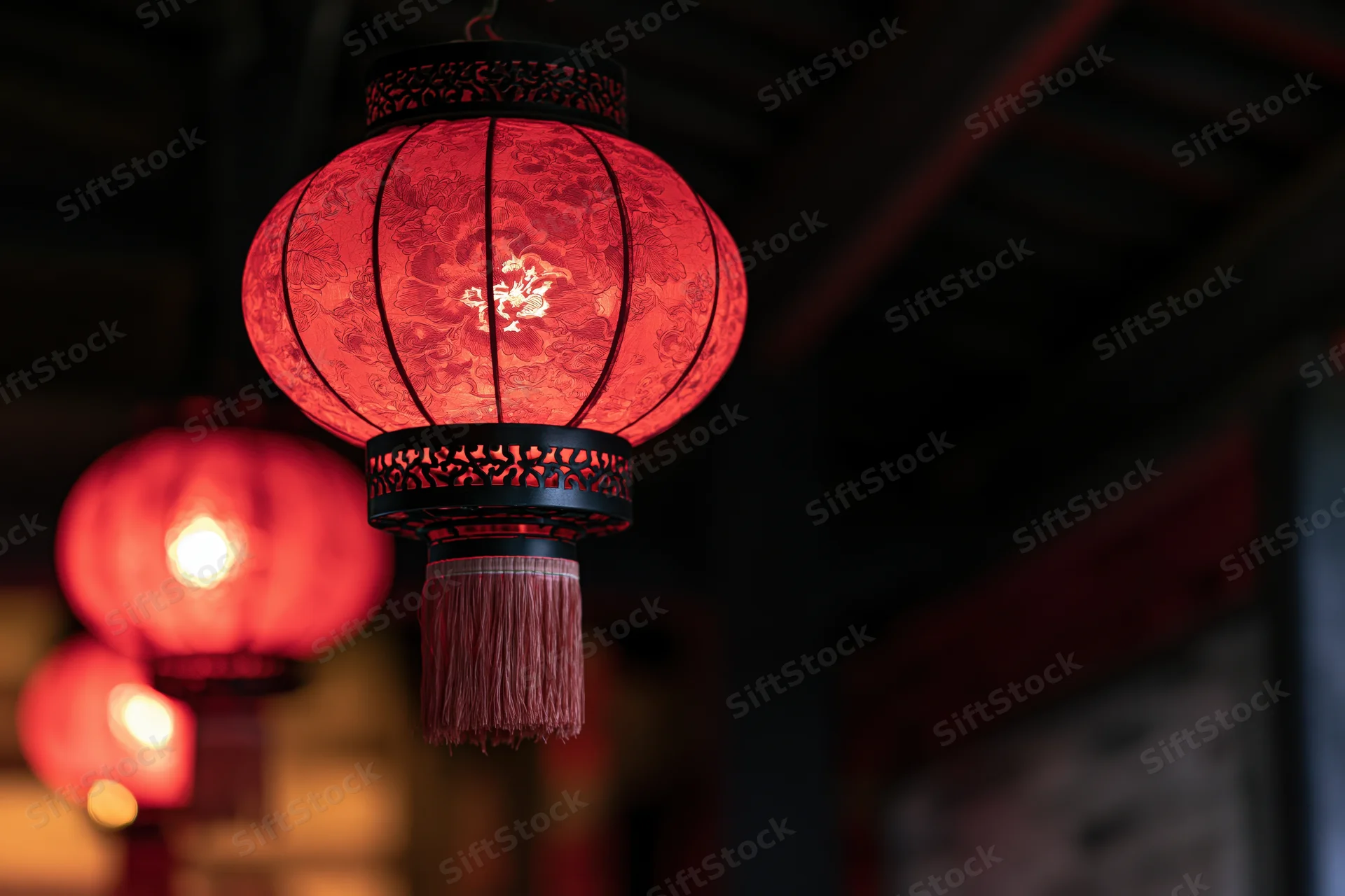 Glowing red chinese paper lantern hanging from dark wooden ceiling with tassel and intricate pattern