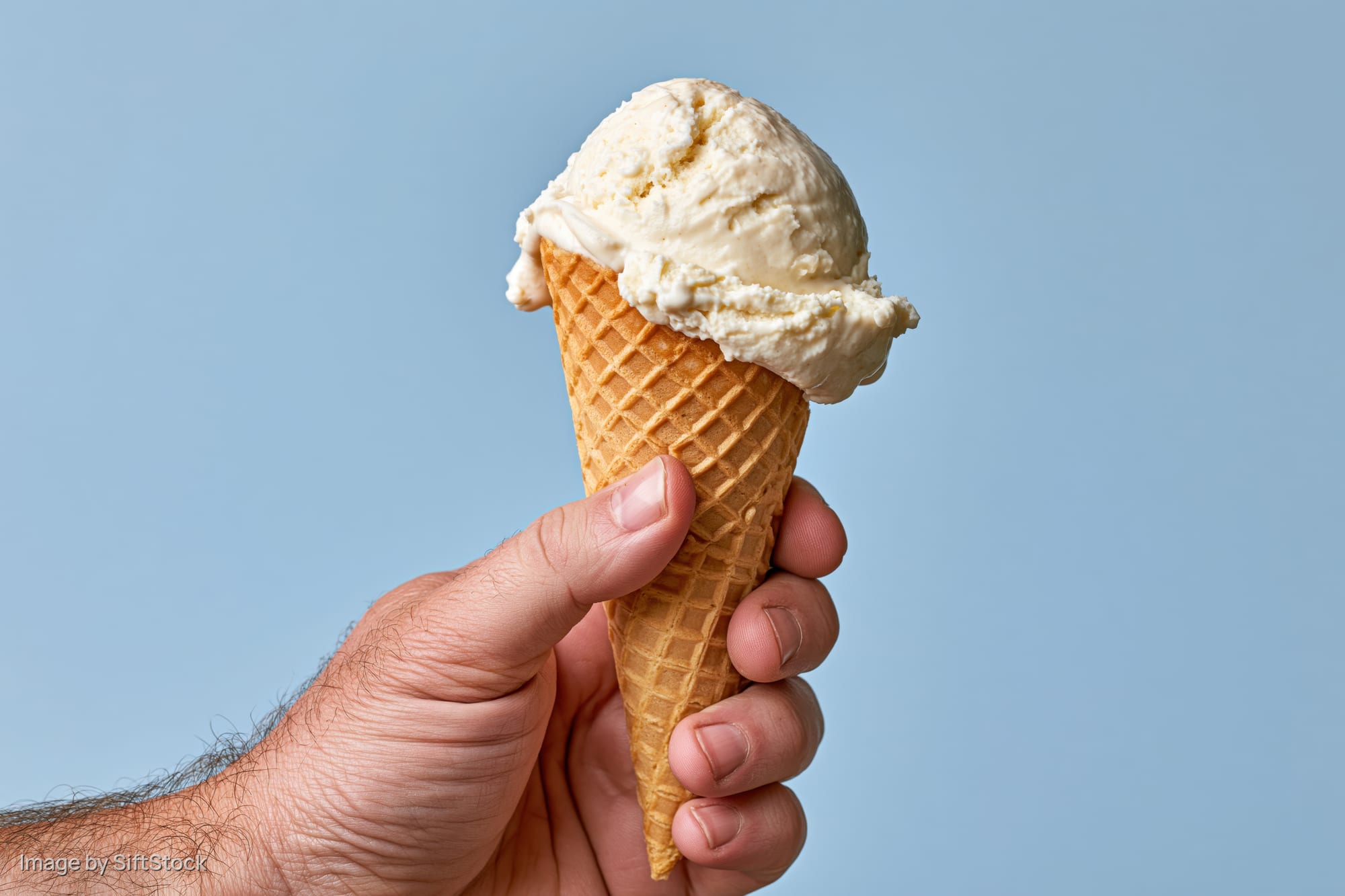 Hand holding a single-scoop vanilla ice cream waffle cone against a light blue background