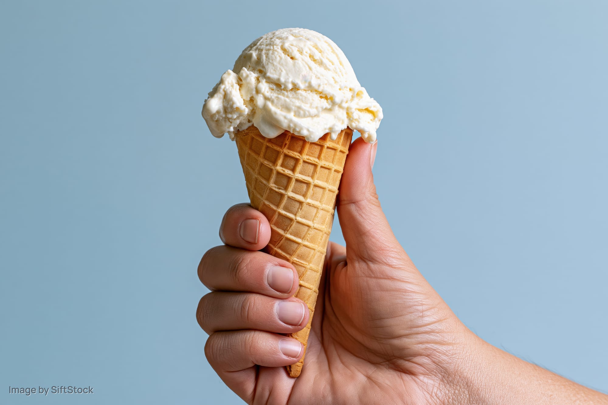 Hand holding a single-scoop vanilla ice cream waffle cone against a light blue background