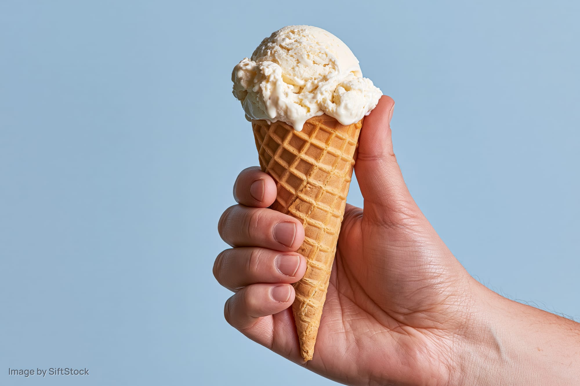 Hand holding a single-scoop vanilla ice cream waffle cone against a light blue background
