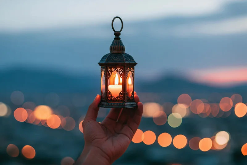 Hand holding ornate metal lantern with lit candle against blurred city lights at twilight