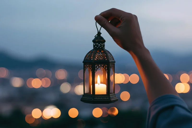 Hand holding ornate metal lantern with lit candle against blurred city lights at twilight