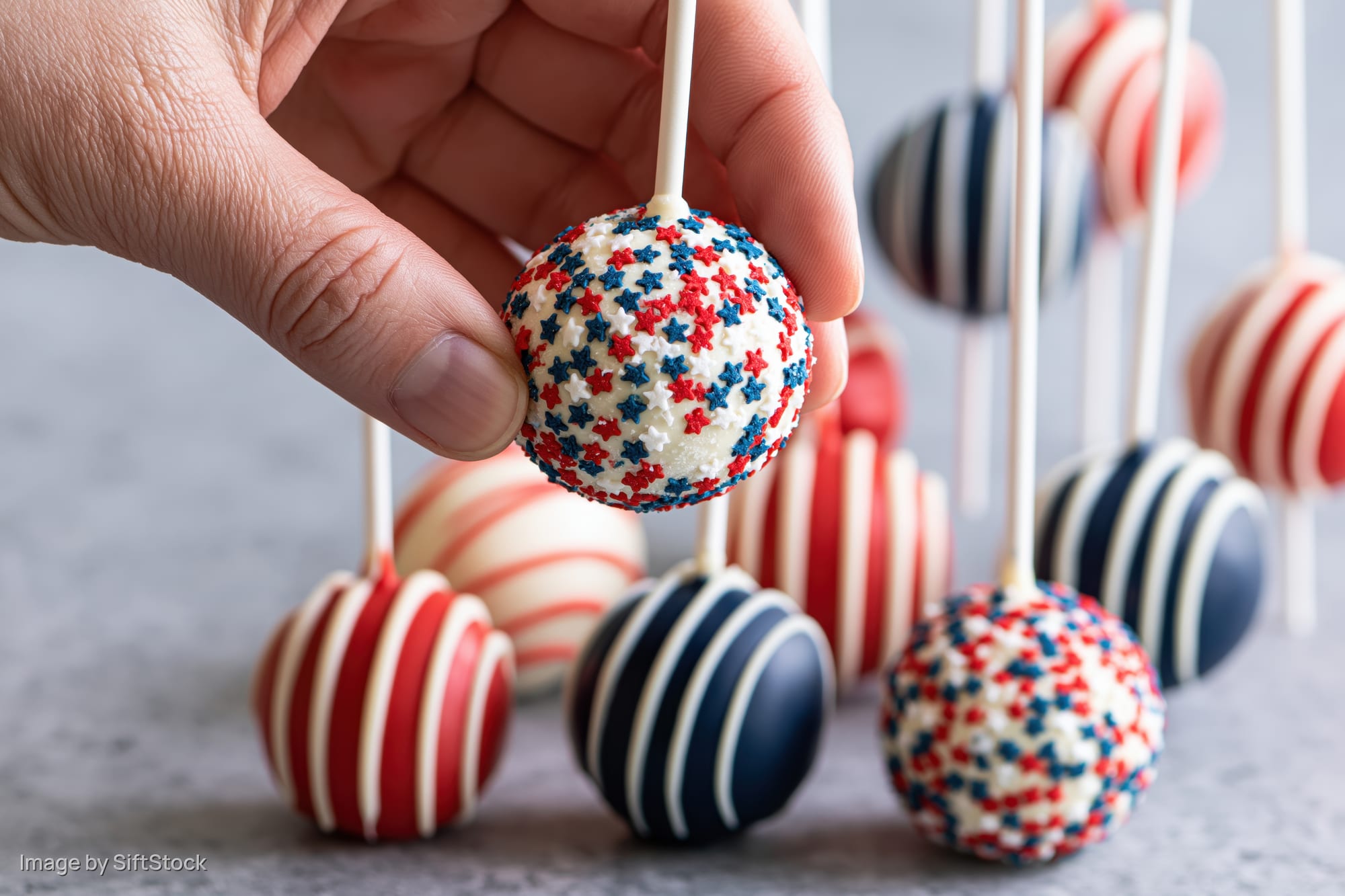 Hand holding a red white and blue star-sprinkled cake pop among striped cake pops on a gray surface