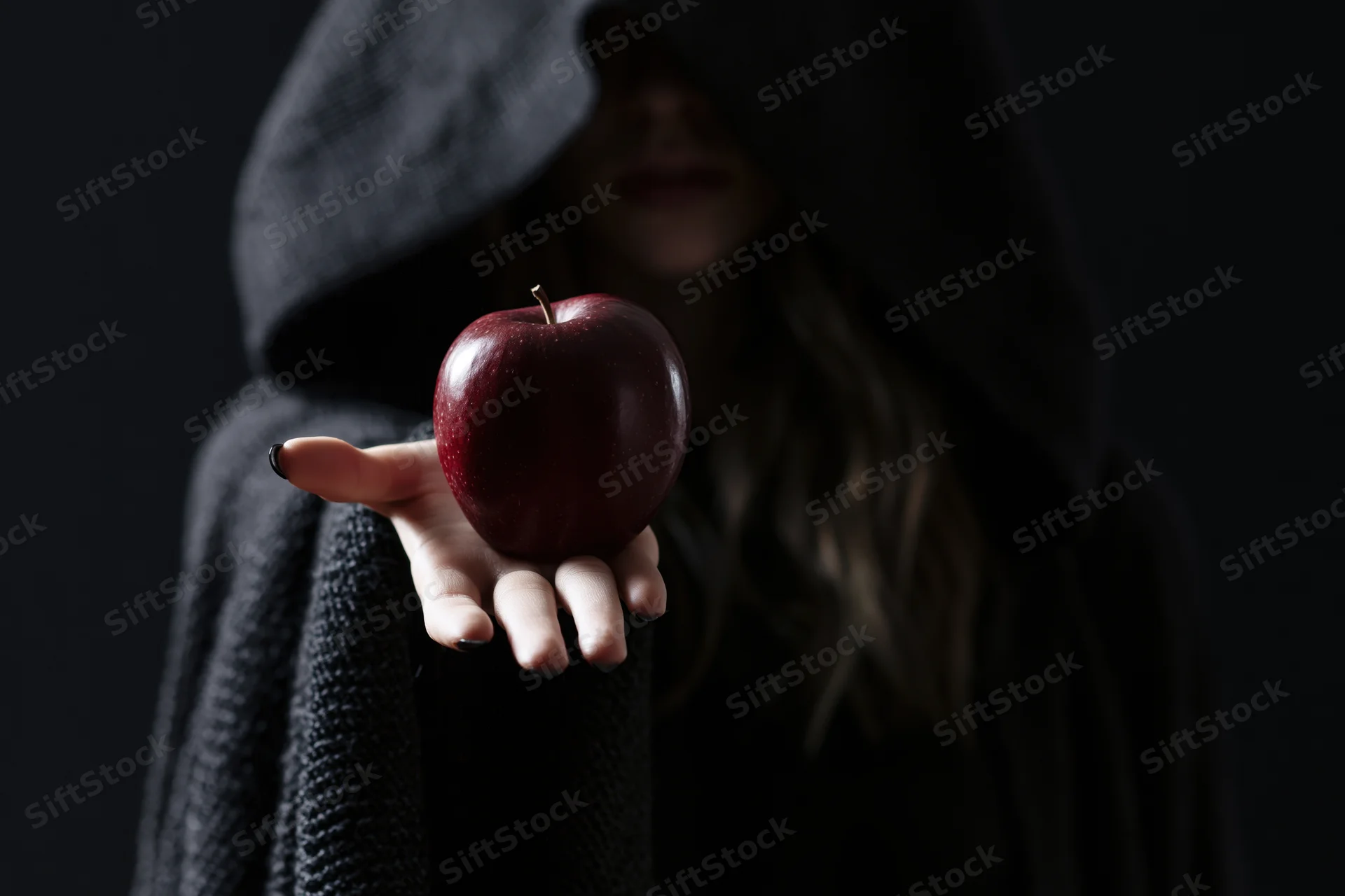 Hooded woman in dark cloak holding red apple in outstretched hand against black background