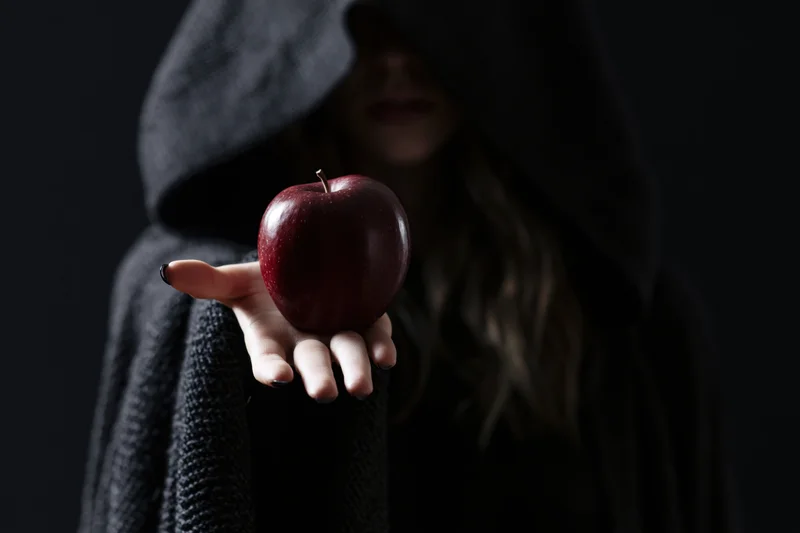 Hooded woman in dark cloak holding red apple in outstretched hand against black background