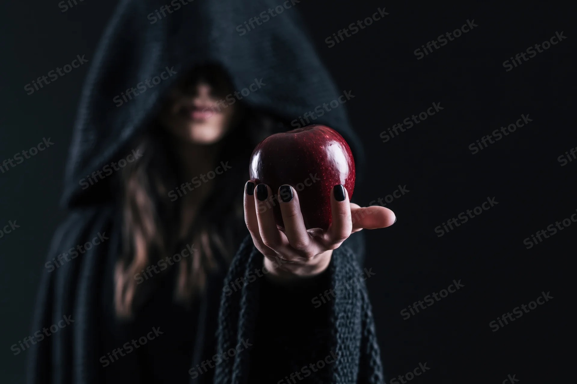 Hooded woman in dark cloak offering red apple with black-polished nails against dark background