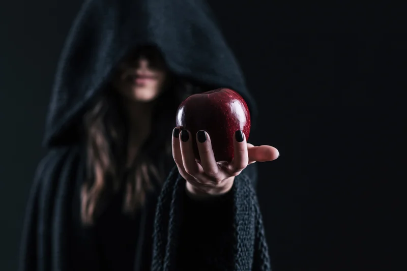 Hooded woman in dark cloak offering red apple with black-polished nails against dark background