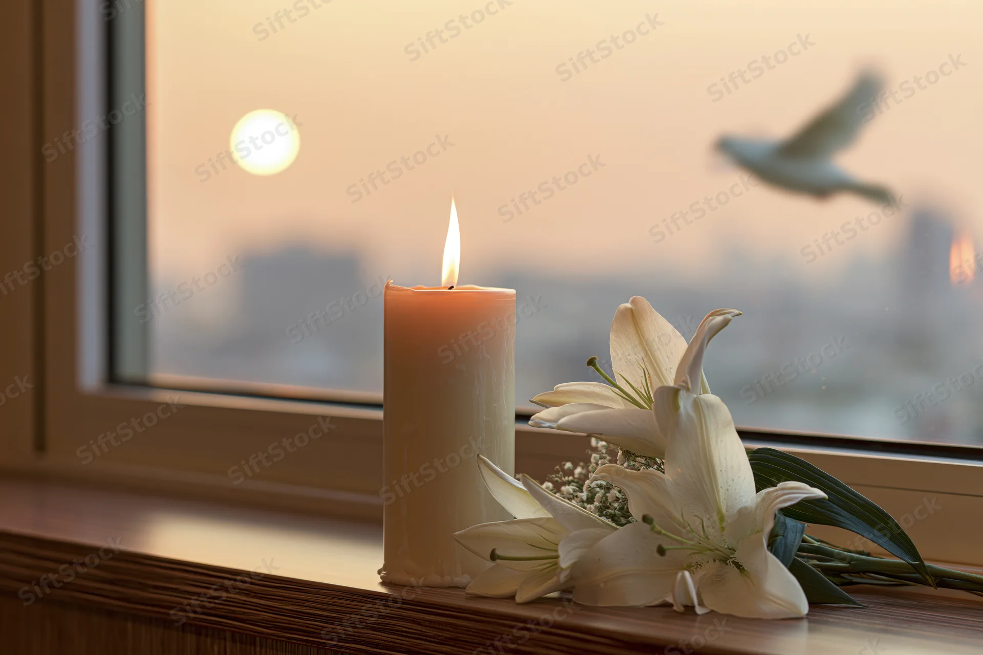Lit white pillar candle and white lilies on a windowsill at sunset