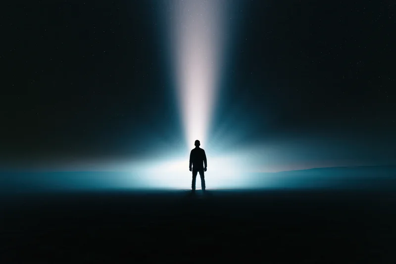 Silhouette of a man standing alone in a dark landscape under a beam of light at night