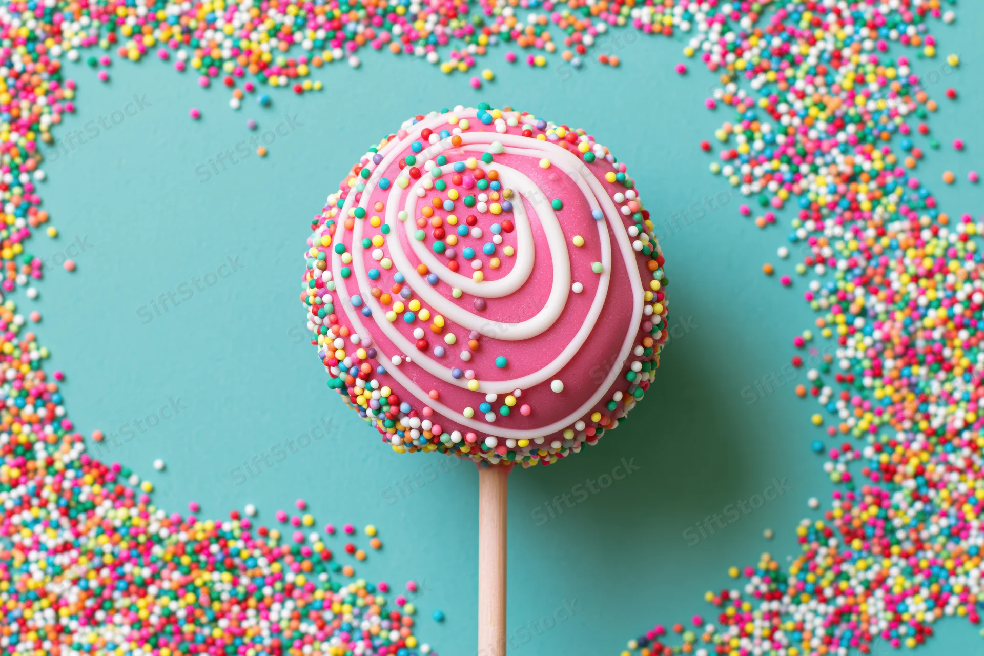 Pink-glazed cake pop with white swirl and colorful sprinkles on wooden stick, surrounded by scattered sprinkles on turquoise surface