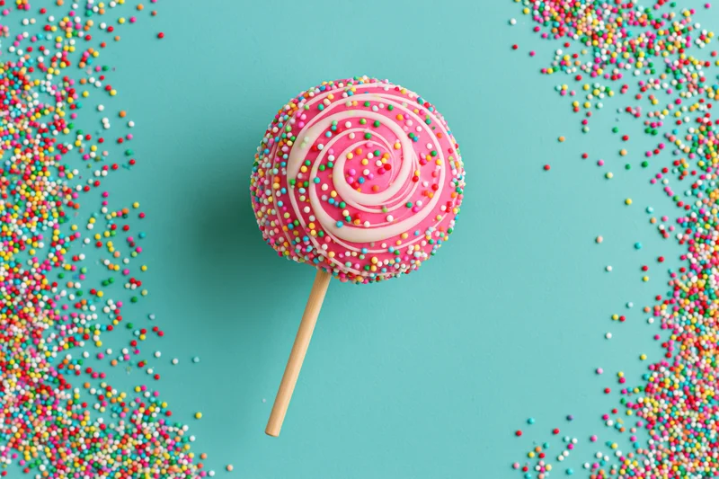 Pink cake pop with colorful sprinkles on wooden stick against turquoise background