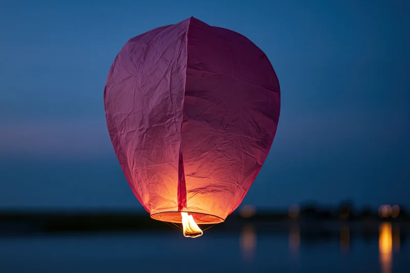 Pink-purple paper sky lantern floating above water at twilight with flame glowing inside