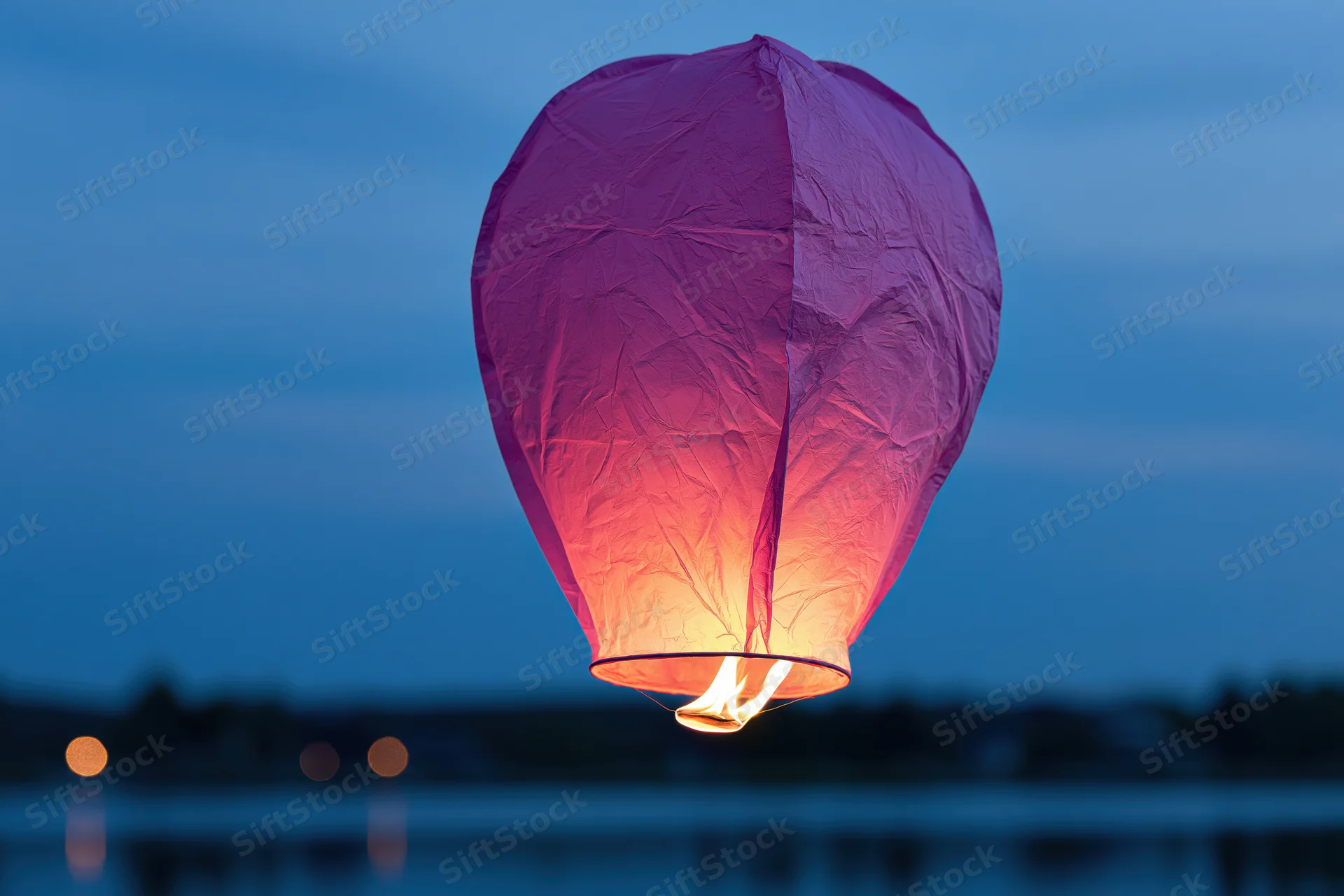 Pink-purple paper sky lantern floating in twilight above water with flame glowing inside