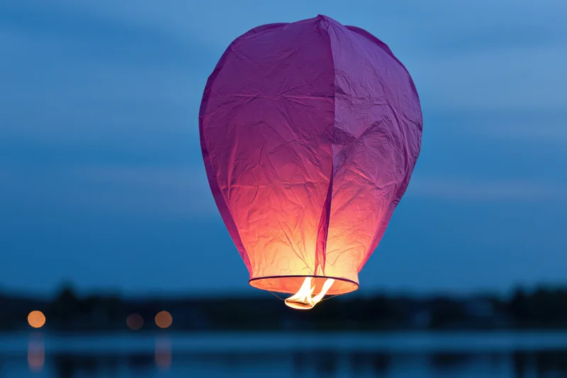 Pink-purple paper sky lantern floating in twilight above water with flame glowing inside
