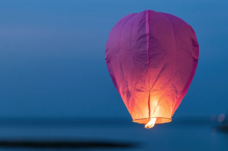 Pink-purple paper sky lantern floating in twilight sky with flame glowing at base