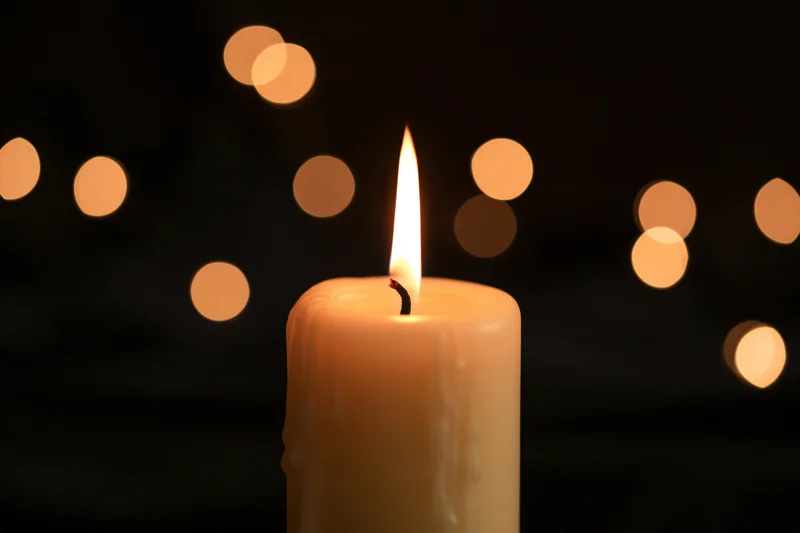 A cream pillar candle burning with a steady warm flame against a dark background with blurred lights