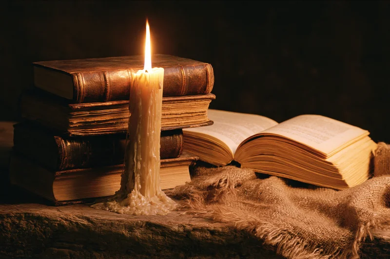 Lit candle beside a stack of old books on a wooden surface with burlap cloth