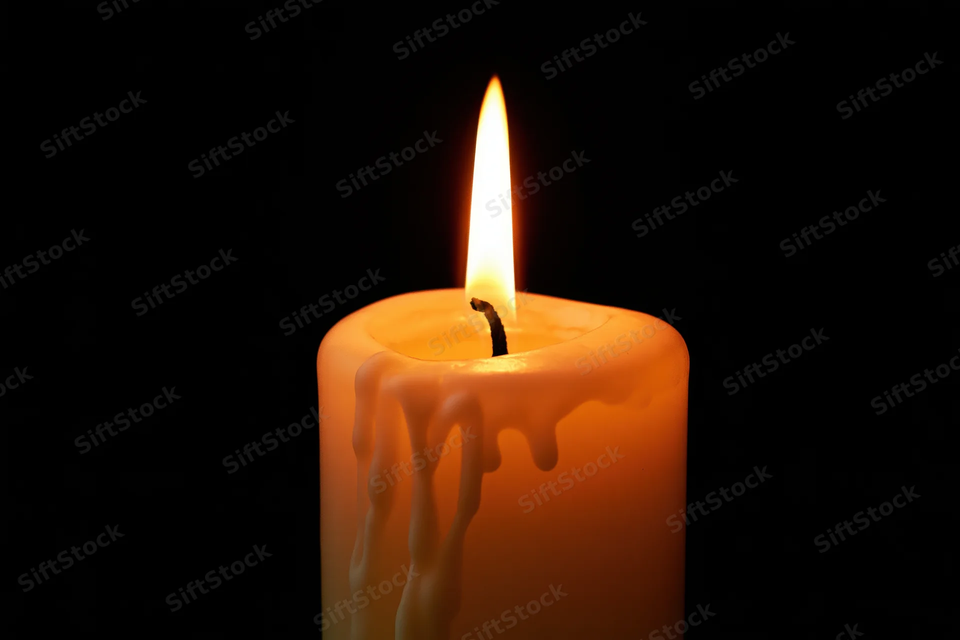 A lit ivory pillar candle with melted wax dripping down its side against a black background