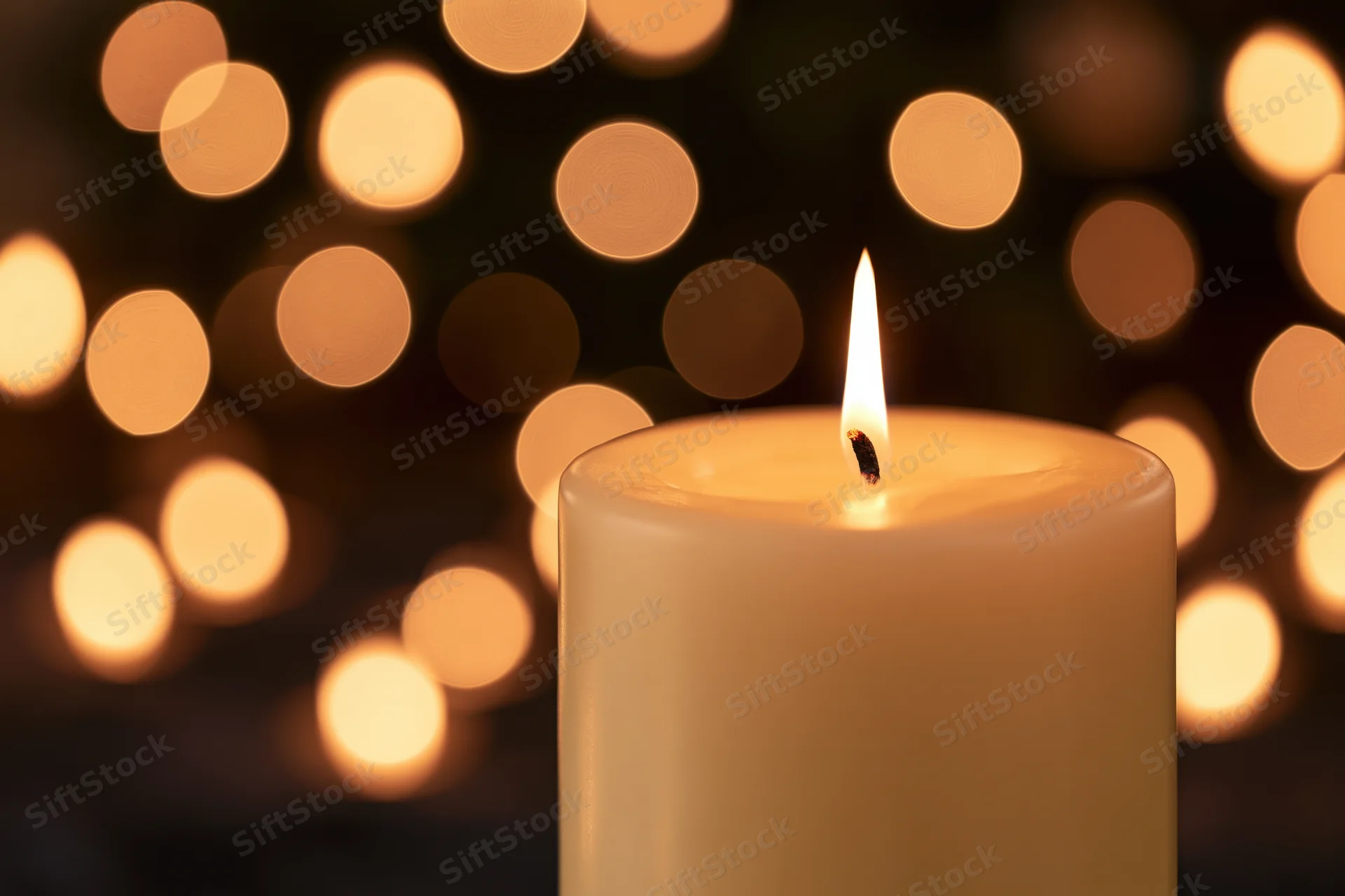 A single white pillar candle burning with a steady flame in front of blurred warm lights
