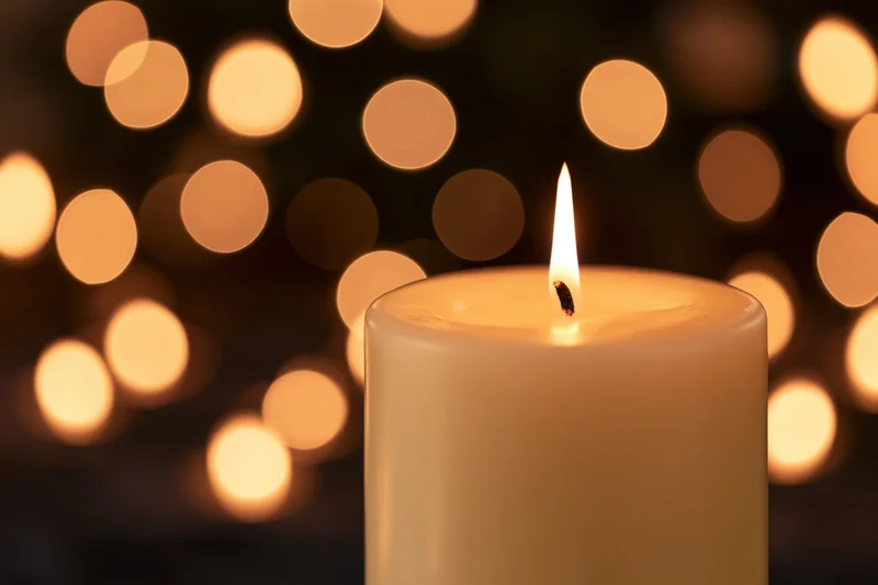 A single white pillar candle burning with a steady flame in front of blurred warm lights
