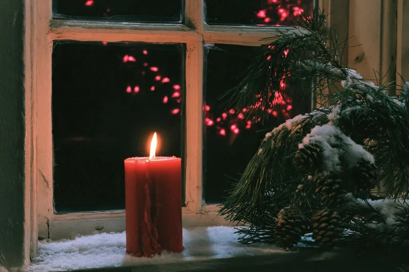 Red candle burning on snow-dusted windowsill beside pine branches and pinecones at night