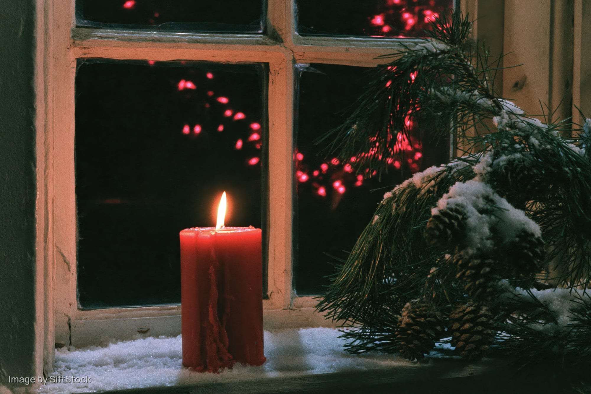 Red candle burning on snow-dusted windowsill beside pine branches and pinecones at night