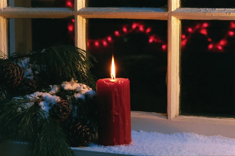 Red candle burning on snow-dusted windowsill beside pine branches and pinecones at night