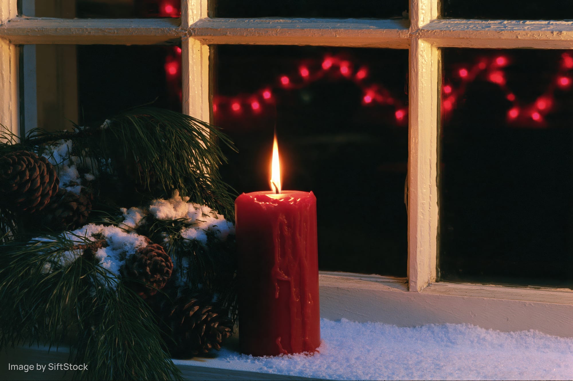 Red candle burning on snow-dusted windowsill beside pine branches and pinecones at night