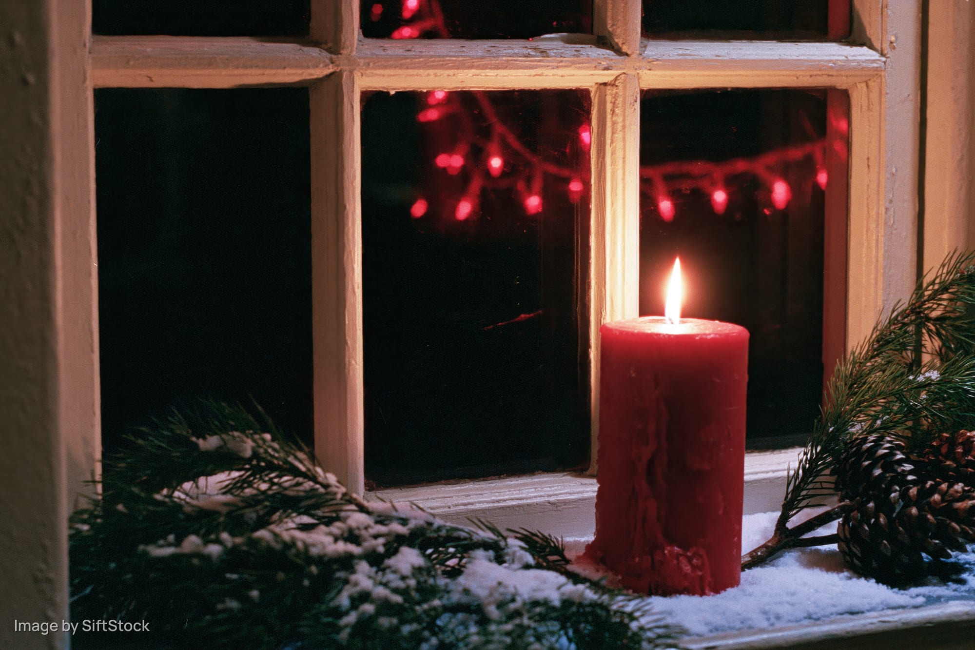 Red candle burning on snow-dusted windowsill with pine branches and pinecone, reflecting red string lights outside at night