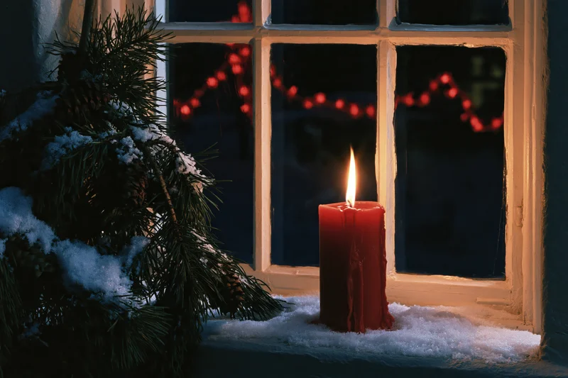Red candle burning on snow-dusted windowsill beside pine branch at night