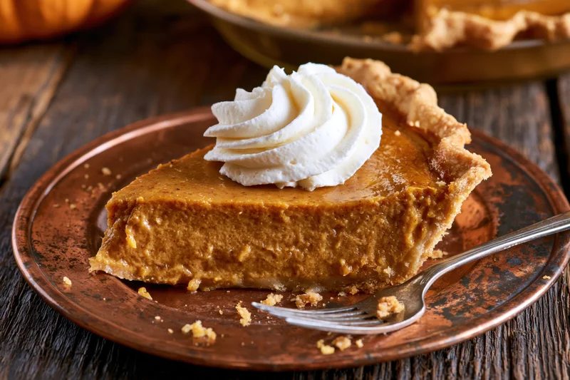 Slice of pumpkin pie with whipped cream on a copper plate with fork on wooden table