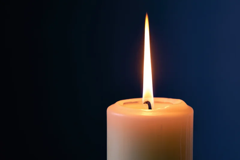 White pillar candle burning with a tall steady flame against dark blue background