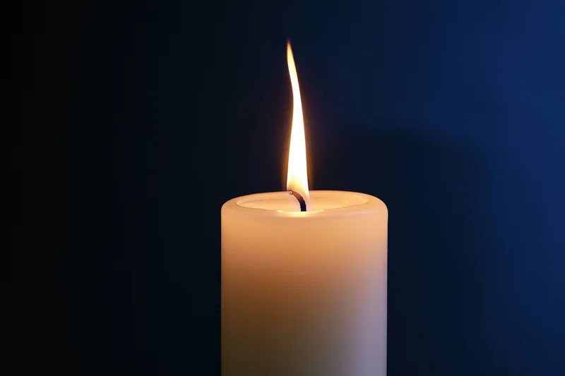 Single white pillar candle burning with a tall steady flame against a dark blue background