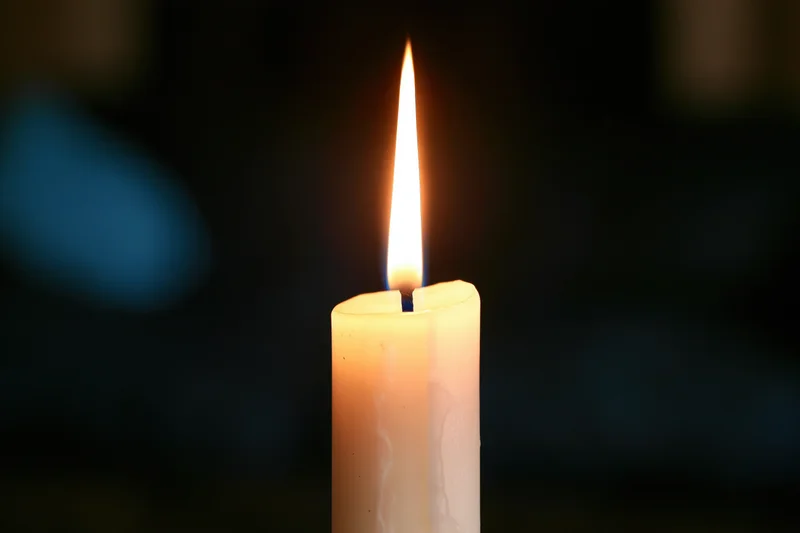 A white taper candle burning with a steady warm flame in a dark setting