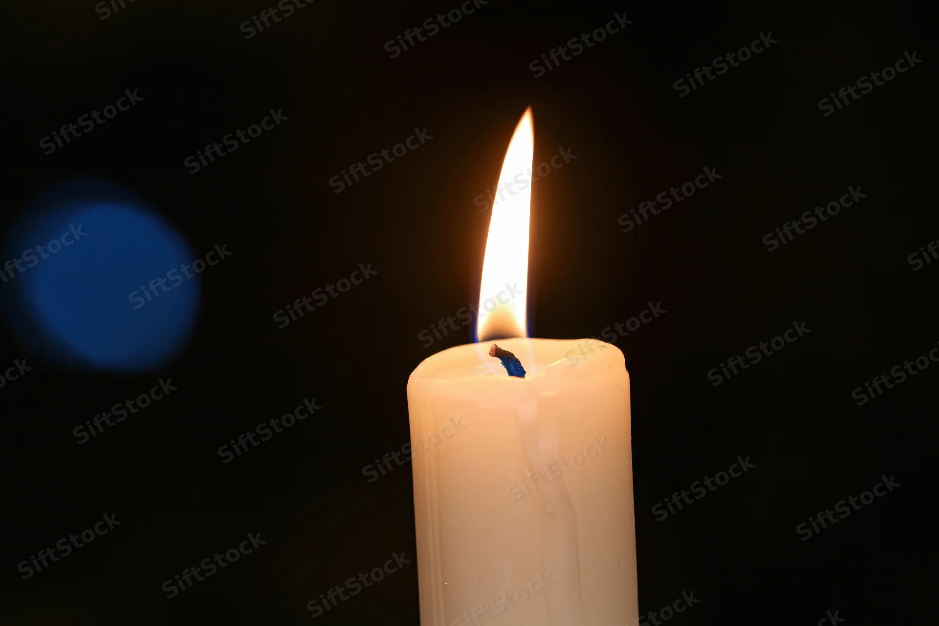 A white taper candle burning with a steady warm flame against a dark background with a blue bokeh light