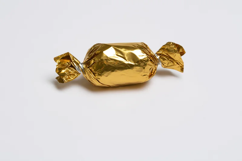Single candy wrapped in crinkled gold foil with twisted ends on white background