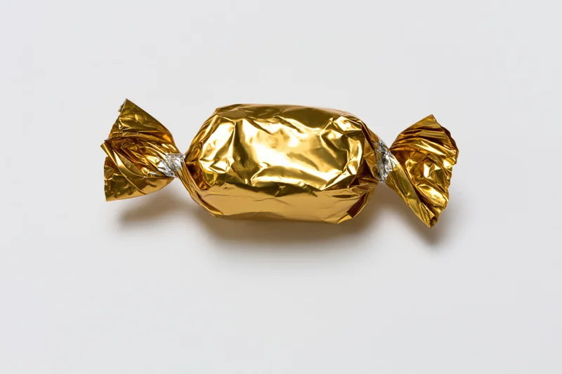 Single candy wrapped in crinkled gold foil with twisted ends on white background
