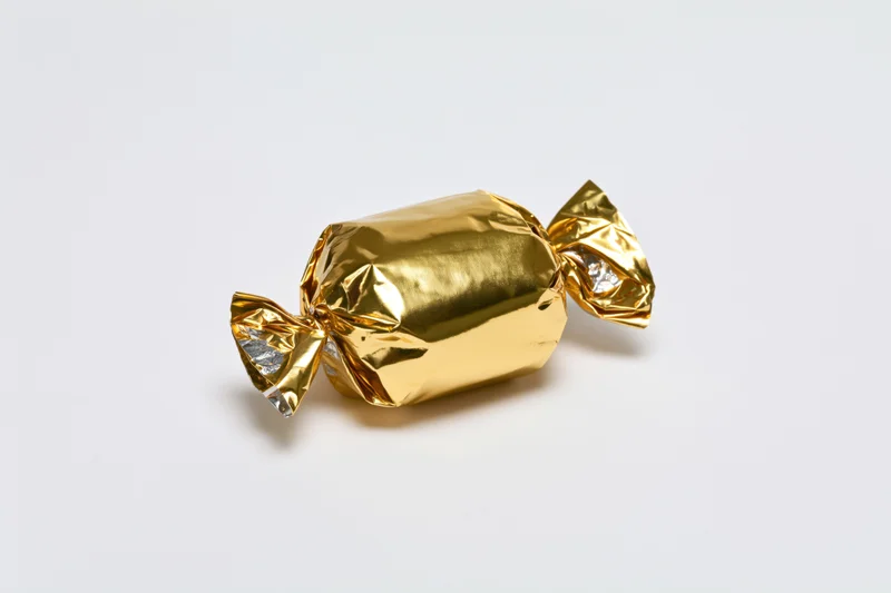 Single candy wrapped in crinkled gold foil with twisted ends on white background