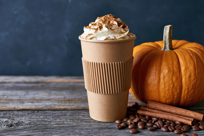 Tall kraft paper coffee cup with whipped cream and cinnamon next to a pumpkin on a wooden table