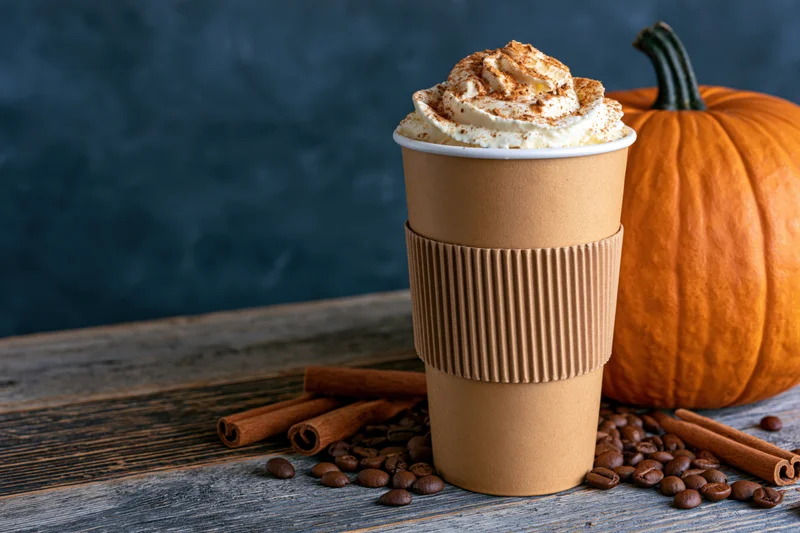 Pumpkin spice latte in a kraft paper cup with whipped cream and cinnamon on a wooden table beside a pumpkin and coffee beans