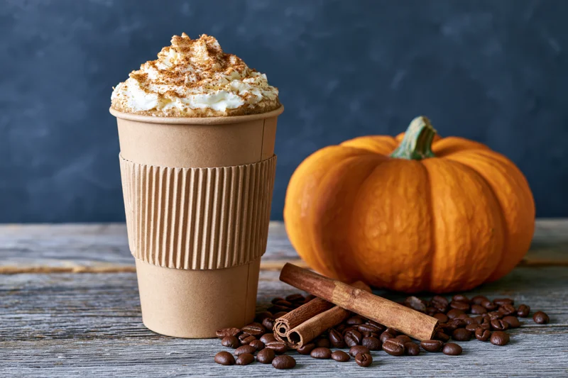 Tall kraft paper coffee cup with whipped cream and cinnamon topping beside a pumpkin and coffee beans on a wooden table