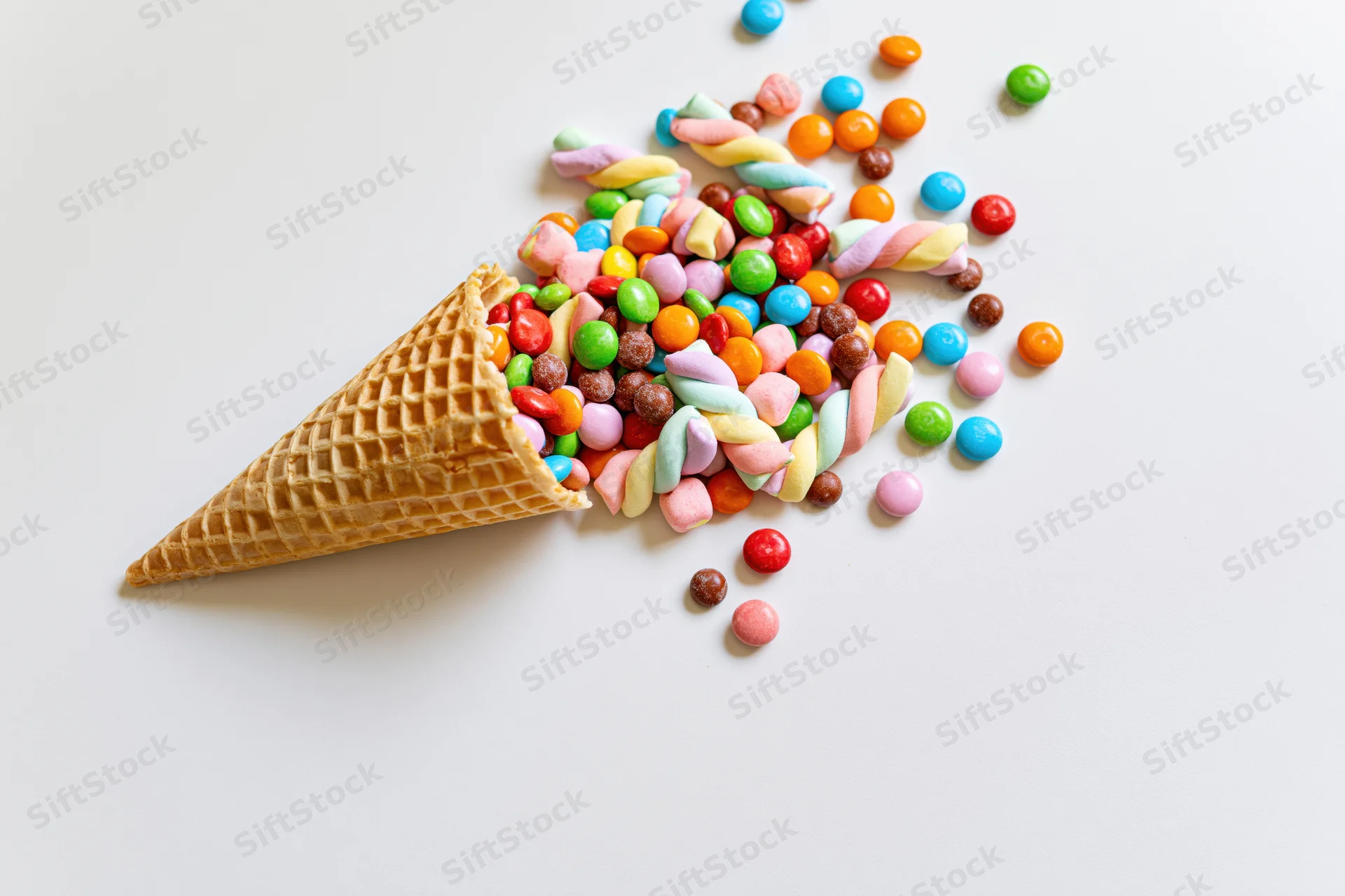 Waffle ice cream cone lying on its side with colorful candies spilled out onto white surface