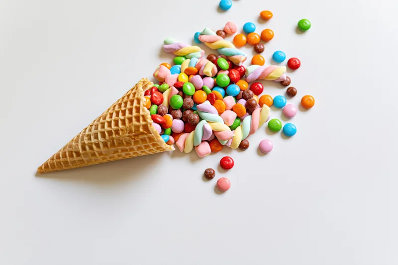 Waffle ice cream cone lying on its side with colorful candies spilled out onto white surface