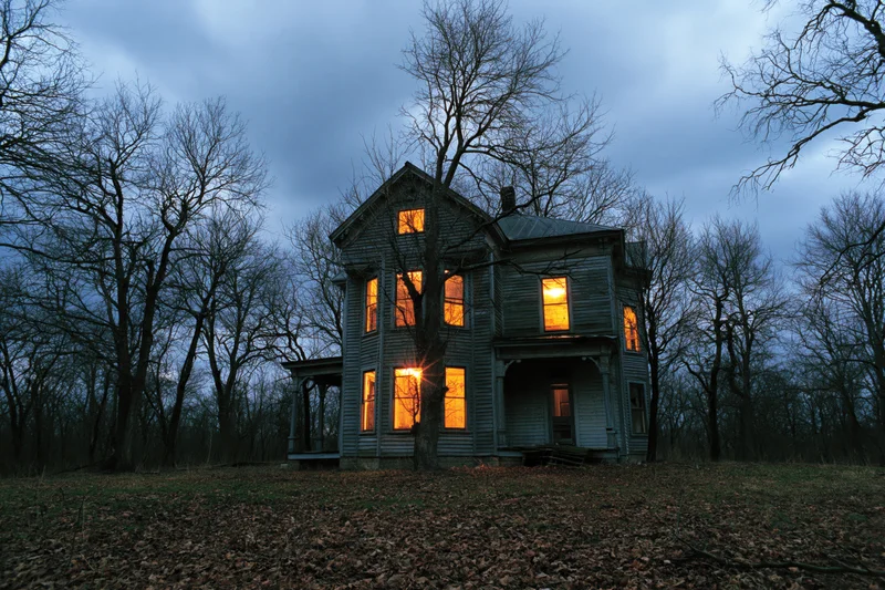 Abandoned victorian house with glowing amber windows at twilight in a leafless forest