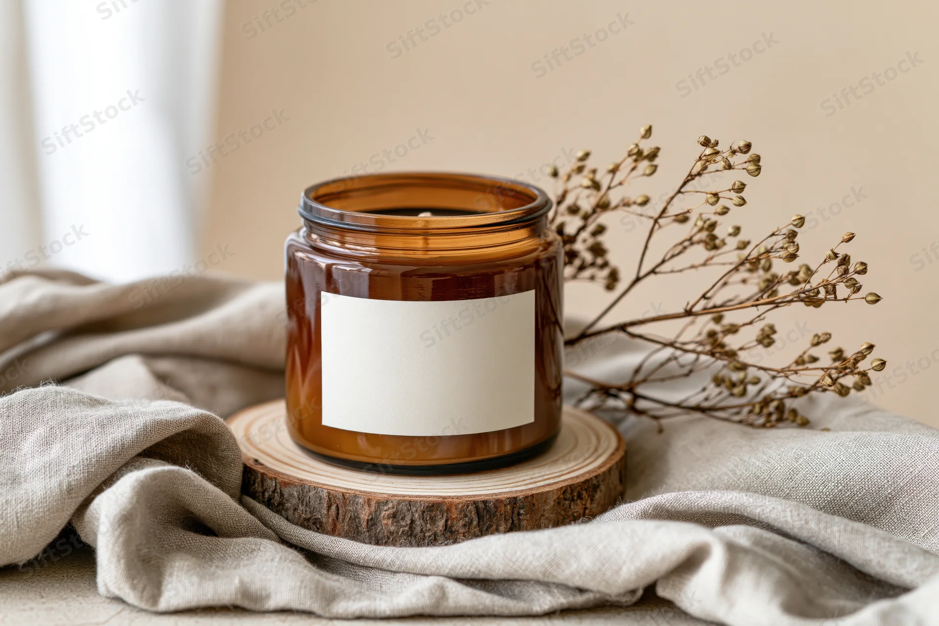 Amber glass candle jar with blank white label on wooden slice beside dried flowers on linen fabric