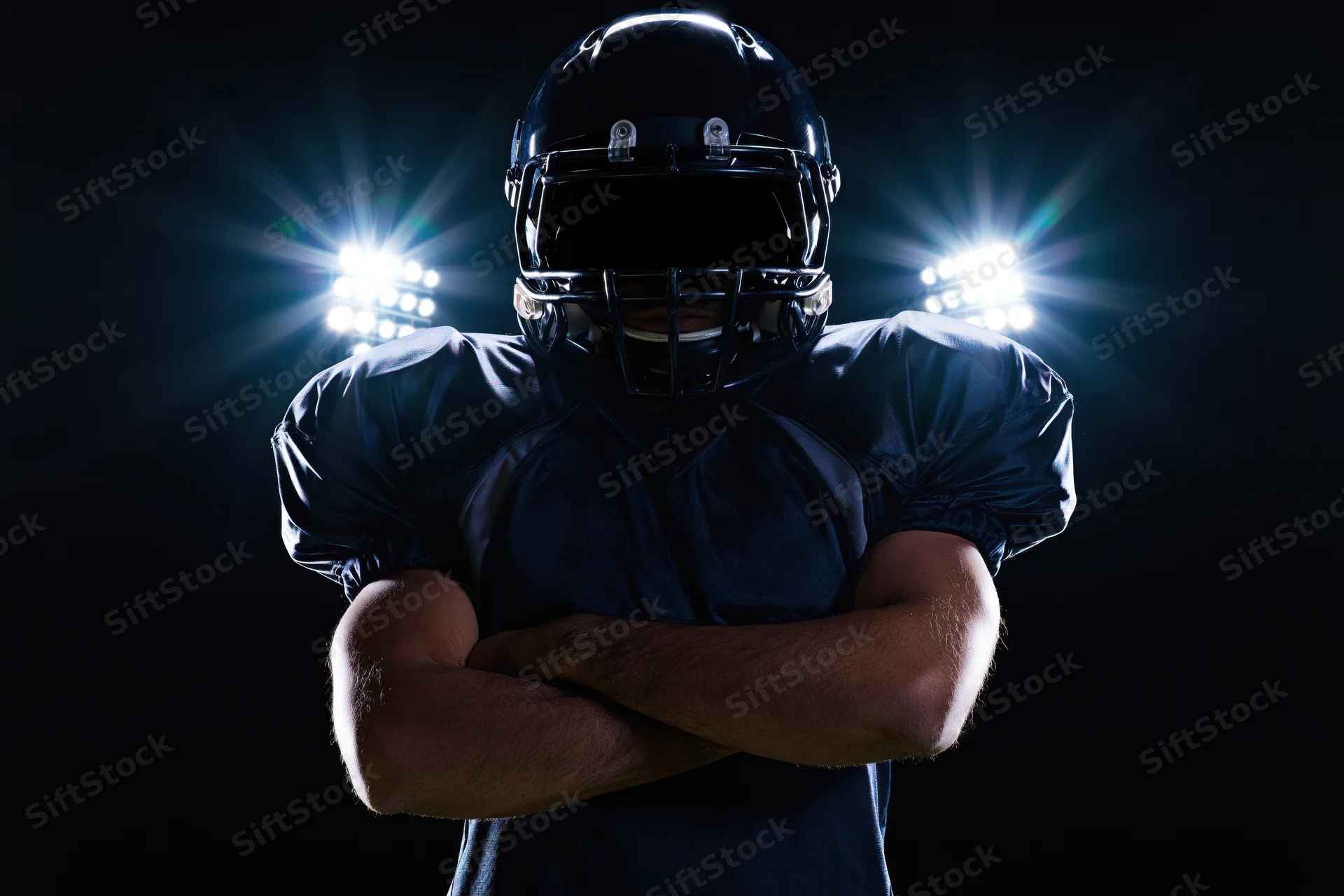 American football player standing with arms crossed facing camera in dark stadium under spotlights