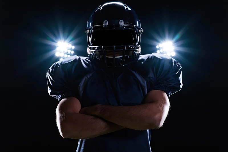American football player standing with arms crossed facing camera in dark stadium under spotlights