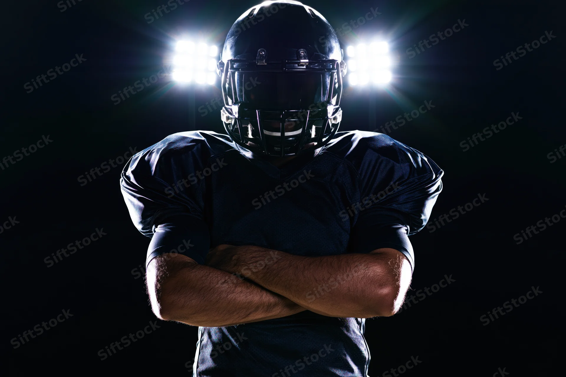 American football player standing with arms crossed in dark setting under spotlight