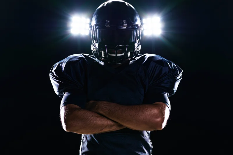 American football player standing with arms crossed in dark setting under spotlight